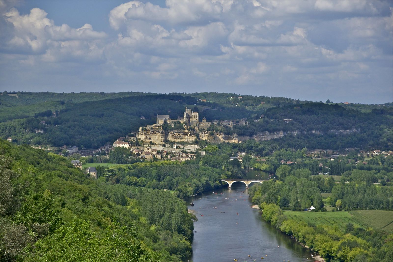 View along the Dordogne valley from Castelnaud: the river curving under a stone bridge, with canoes on the water, towards the Château de Beynac on its cliff above the village, framed by wooded hills.