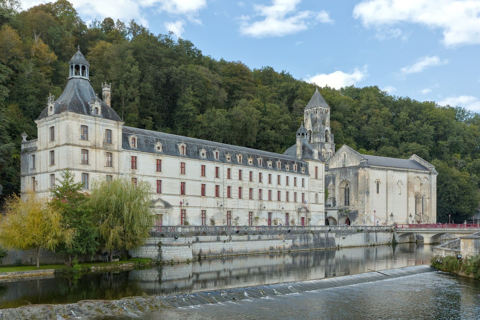 The long pale stone abbey buildings of Brantôme and the church's tall gabled bell tower beside the River Dronne, reflected in the water above a low weir, with a wooded hillside behind.