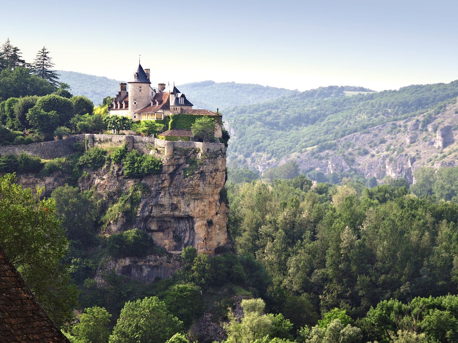 The Château de Belcastel, with its pointed tower and walled terrace, perched on the edge of a sheer limestone cliff above wooded slopes, with forested hills and rocky outcrops behind.