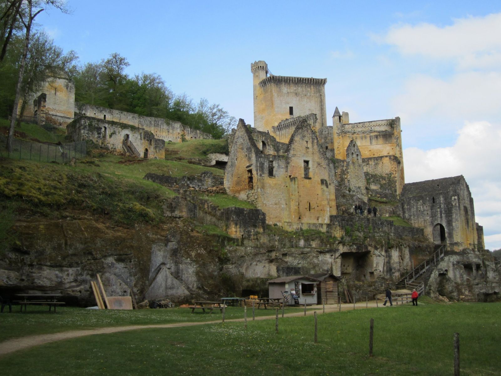 The ruins of the medieval Château de Commarque on a limestone outcrop, with a tall square keep and roofless stone houses above cliff shelters, a grassy meadow and a path in front.