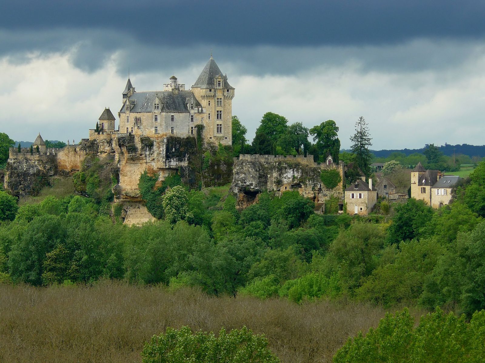 The turreted Château de Montfort perched on a rocky limestone cliff above dense green woodland, with village houses to the right under a dark, stormy sky.