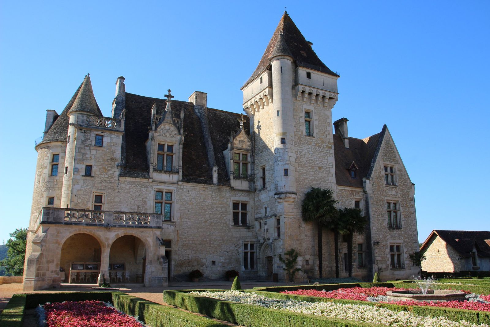 The Renaissance Château des Milandes, with its pointed towers and carved dormer windows, behind a formal garden of clipped box hedges, red and white flower beds and a fountain, under a clear blue sky.