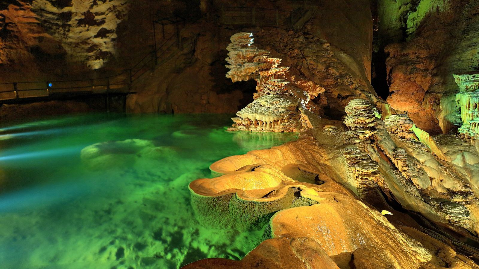 Clear green water of the upper lake inside the Gouffre de Padirac, with lit rimstone pools and tiered calcite formations beside a railed walkway.