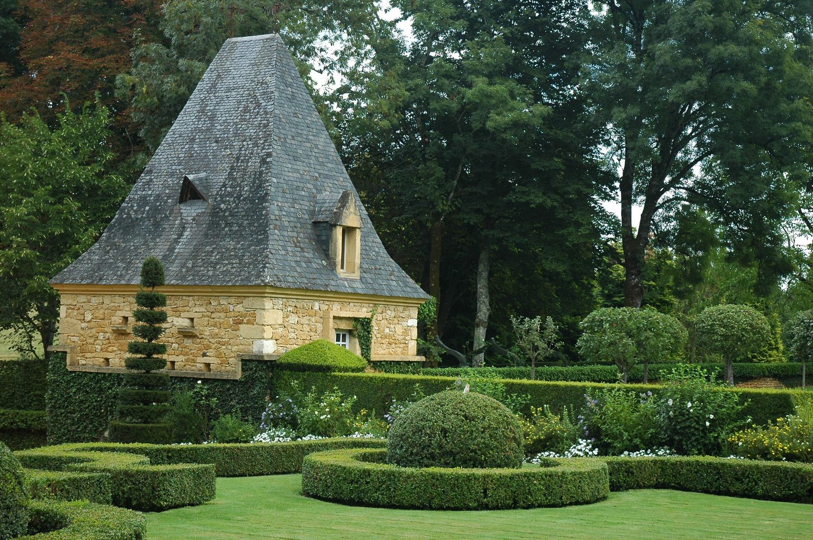 Clipped box hedges, a topiary dome and a tiered spiral of clipped greenery in the Eyrignac gardens, in front of a stone pavilion with a steep pyramid roof and tall trees behind.