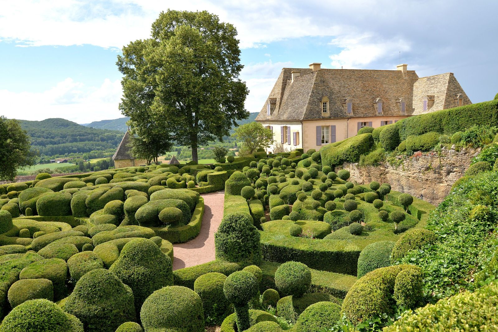 Rounded, cloud-like clipped box hedges and topiary balls in the Jardins de Marqueyssac, with a gravel path, a large tree and the stone-roofed château behind, and wooded hills in the distance.