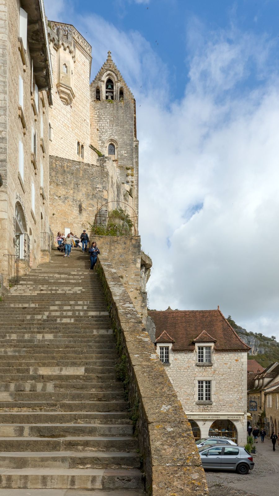 A long, wide flight of worn stone steps climbing beside a parapet towards Rocamadour's religious buildings and a gabled bell tower, with a few visitors near the top, under a blue sky with white cloud.