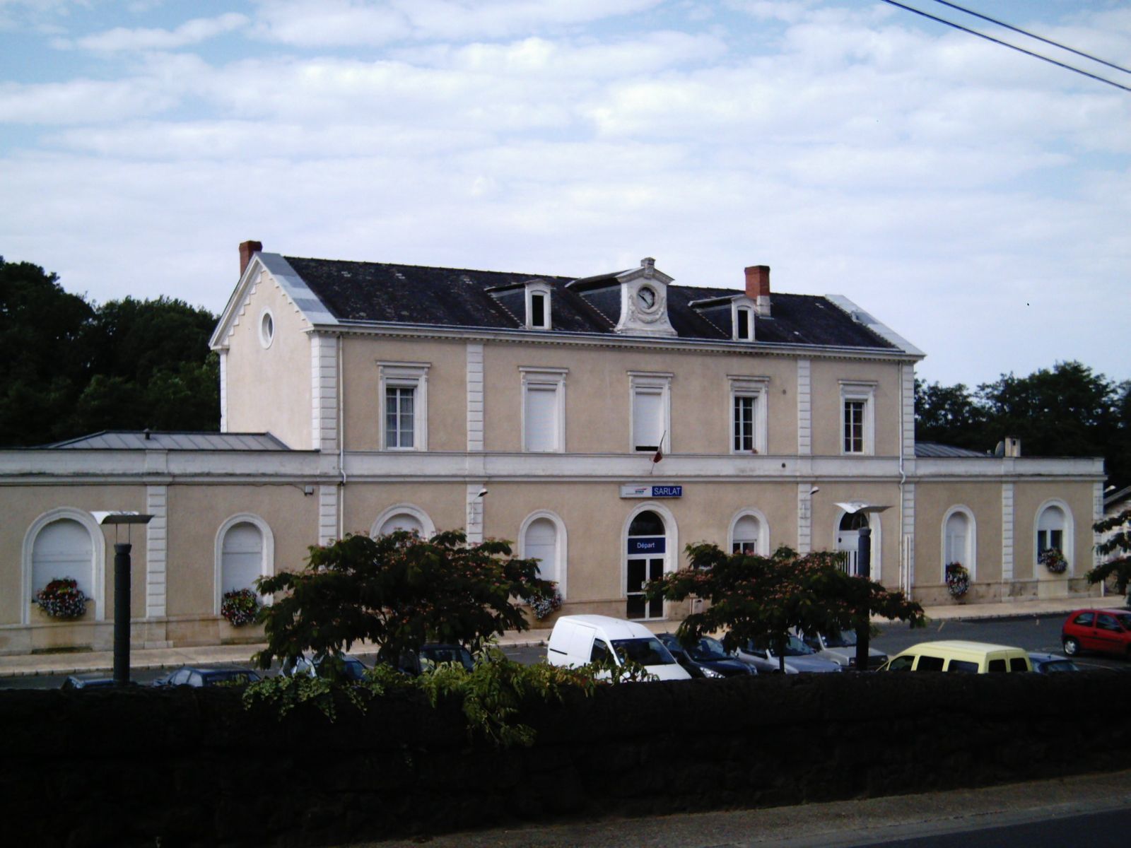 The two-storey cream-coloured station building at Sarlat, with arched ground-floor windows, a small clock on the roof and a SARLAT sign over the door, with parked cars and trees in front.