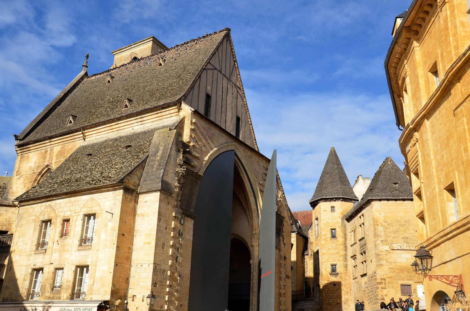 The former Sainte-Marie church in Sarlat, its stone-tiled roof and gable above a tall arched opening fitted with giant dark metal doors, beside honey-coloured houses with pointed stone towers.
