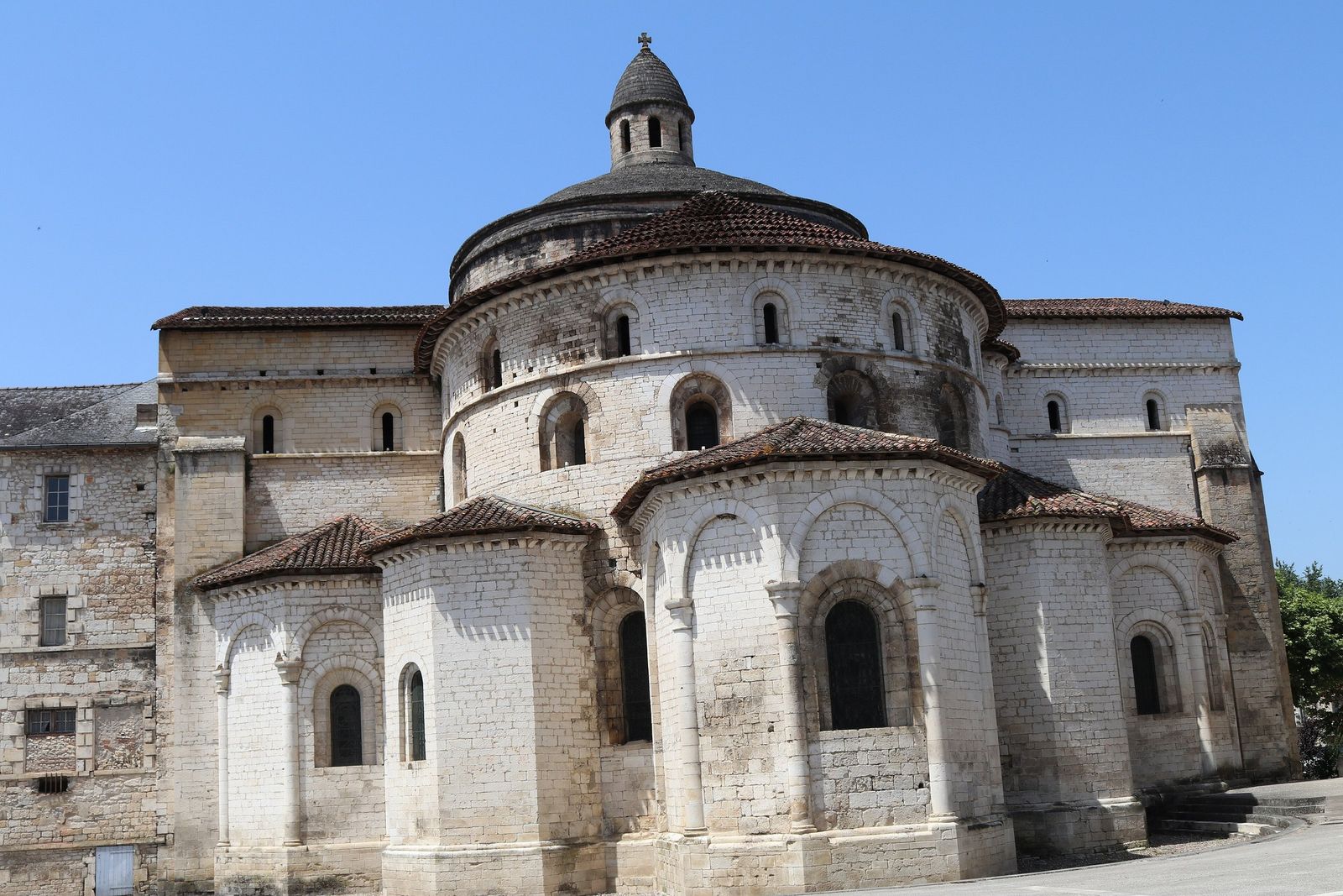 The pale stone east end of Souillac's Romanesque abbey church, with rounded apses and chapels under red-tiled roofs, rising to a dome topped by a small lantern, against a clear blue sky.