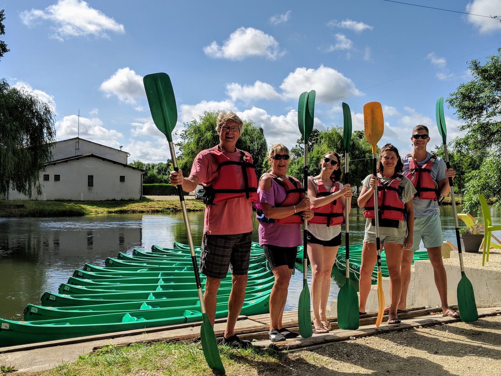 The family in life jackets, holding paddles beside a row of green canoes on a riverbank