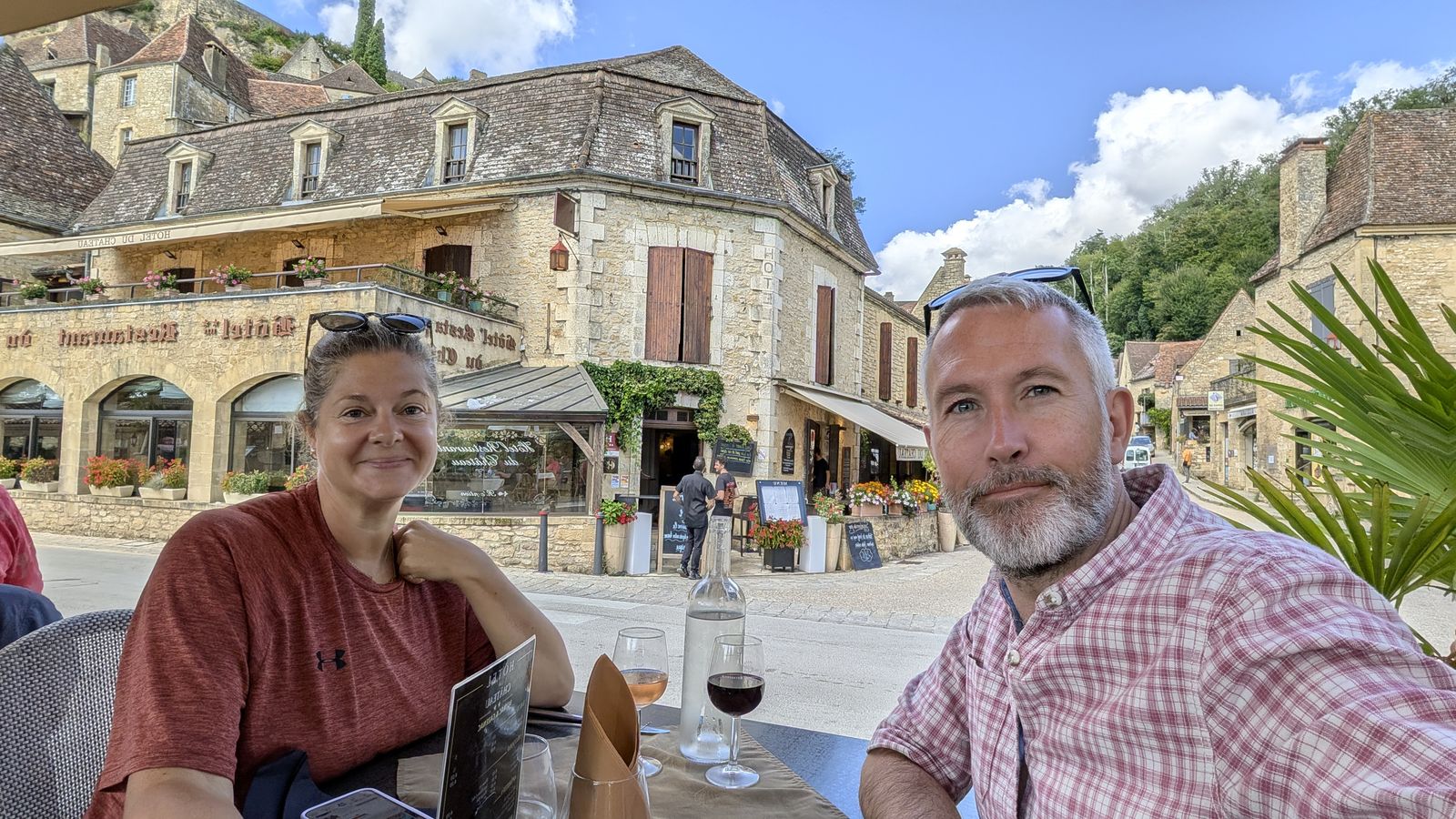 Cassie and Kev with drinks at a café table in Beynac, the village's stone houses behind them