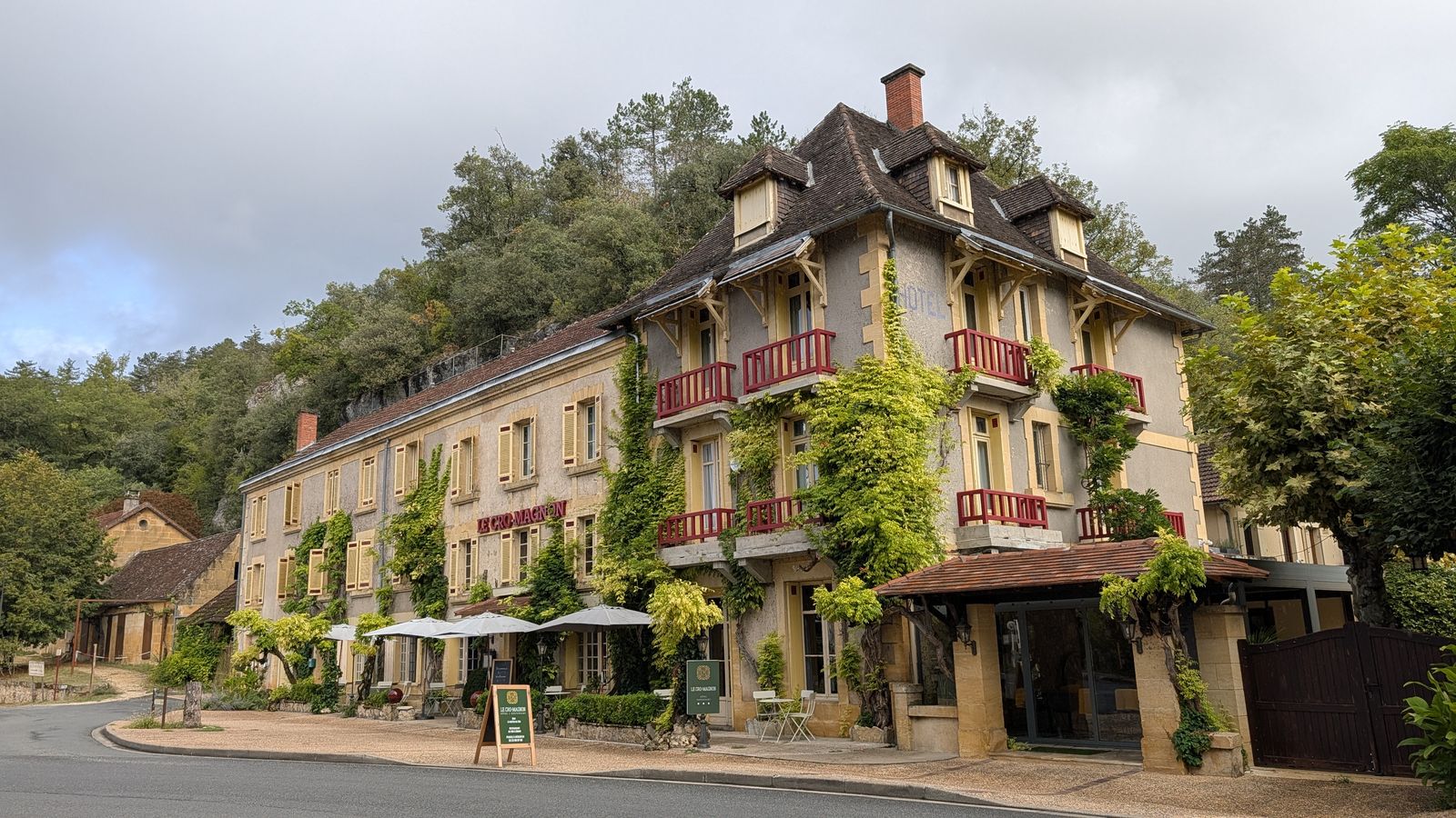 A hotel with balconies and climbing plants beneath a wooded cliff at Les Eyzies