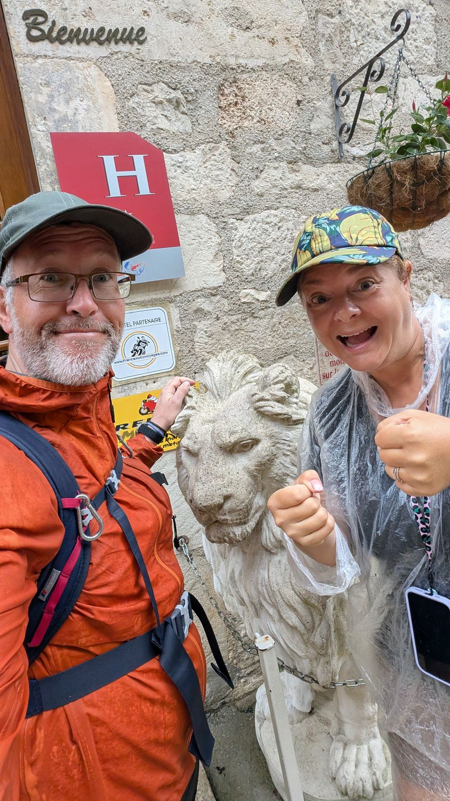 Kev and Cassie in rain gear beside a stone lion at their hotel in Rocamadour