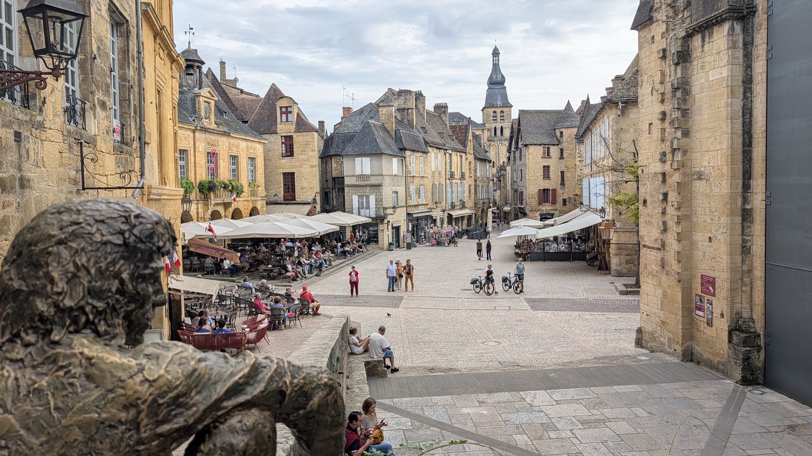A bronze figure on the steps looking down over Place de la Liberté and its café terraces, Sarlat