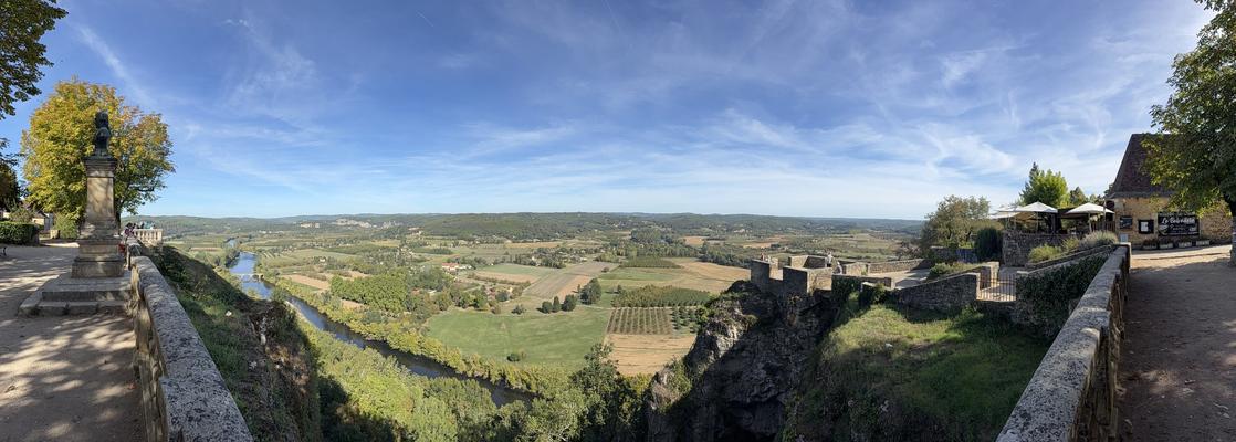 A wide panorama from a clifftop terrace over a river winding through a patchwork of fields