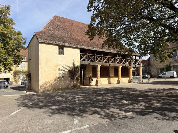 A golden-stone market hall with a wooden balcony on stone pillars, beside a sunny square
