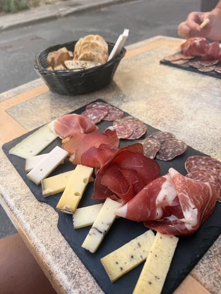 A slate board of cured ham, sausage and cheese on an outdoor table, with a basket of toasted bread