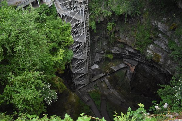 Looking down from the rim into the Gouffre de Padirac, a deep round chasm with green, overgrown rock walls, a tall metal stair and lift tower and walkways at the bottom.