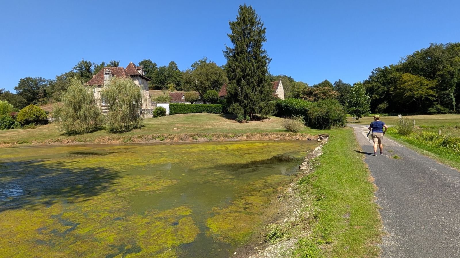 A walker on a sunny country lane past a pond and an old stone manor house